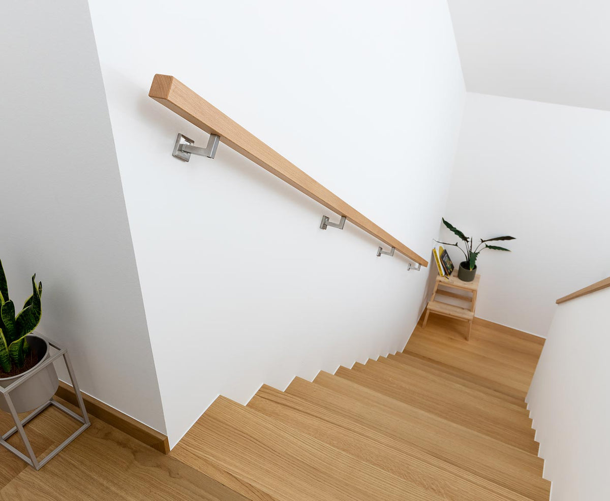 Treppenabgang aus Holz mit langem, quadratischen Handlauf aus Buche, durch Edelstahlhalter an weißer Wand befestigt.