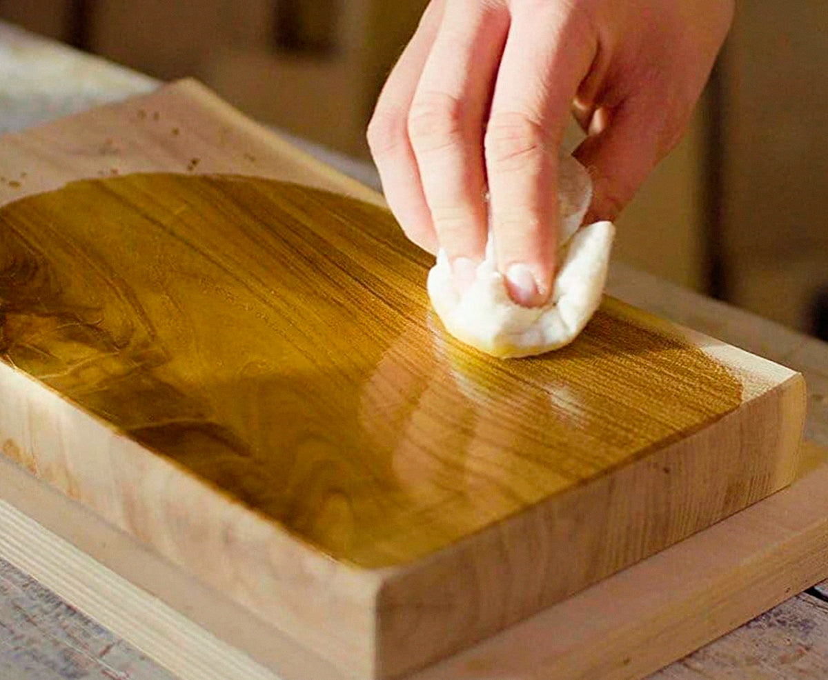 Hand mit weißem Pad, die ein Holzbrett einölt.